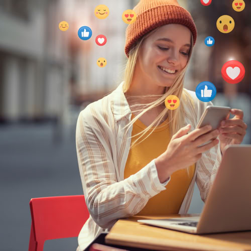 Woman smiling at a phone outdoors with social media reaction icons, laptop on the table.