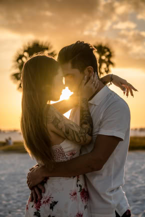 A couple embracing on a beach at sunset, with palm trees in the background.