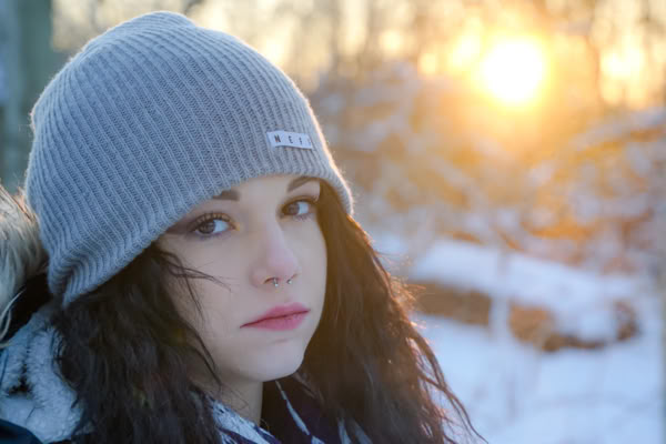 A person wearing a gray beanie and a winter coat stands in a snowy landscape with the sun setting in the background.