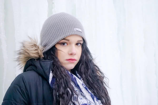 A person wearing a gray beanie and a black coat with a fur-trimmed hood stands in front of an icy background.