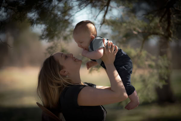 A woman is holding a baby up in the air, both facing each other, with trees and a blurred background.