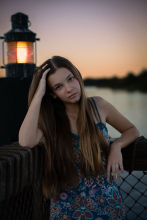 A person with long hair leans on a railing near a lit lantern, with a body of water and a sunset in the background.